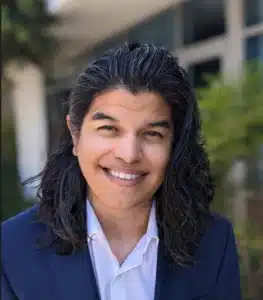 A person with long dark hair wears a navy blue blazer and a white shirt, smiling outdoors in front of a blurred company building and greenery.