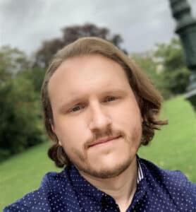 A person with medium-length hair and a light beard, wearing a navy blue polka dot shirt with the company logo, standing outdoors with greenery in the background.
