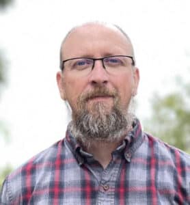 A man with a bald head and a gray beard, wearing glasses and a plaid shirt, stands outside with a blurred background, giving the impression of being near his company.