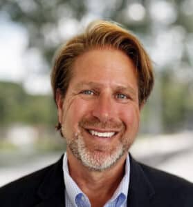 A man with short, light brown hair, a beard, and a friendly smile is dressed in a dark blazer and light blue collared shirt. The out-of-focus background appears to be a park or outdoor setting, giving him a relaxed yet professional air perfect for representing his company.