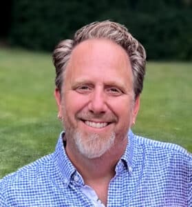 A person with short, light-colored hair and a beard smiles while wearing a patterned shirt, standing outdoors on the green lawn of the company grounds.